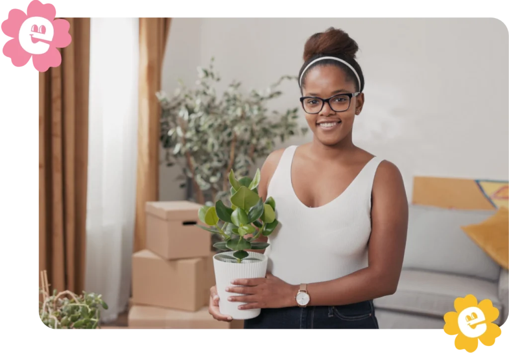A smiling young Black woman stands in her new apartment holding a potted plant. Some boxes are stacked in the background.