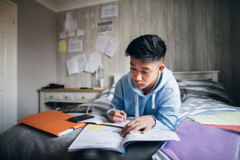 A young Asian man completes his homework on his bed.