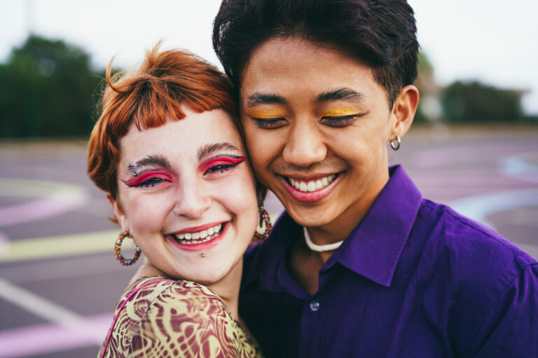 Two smiling young LGBTQ friends standing cheek to cheek. They are both wearing colorful eye make up.