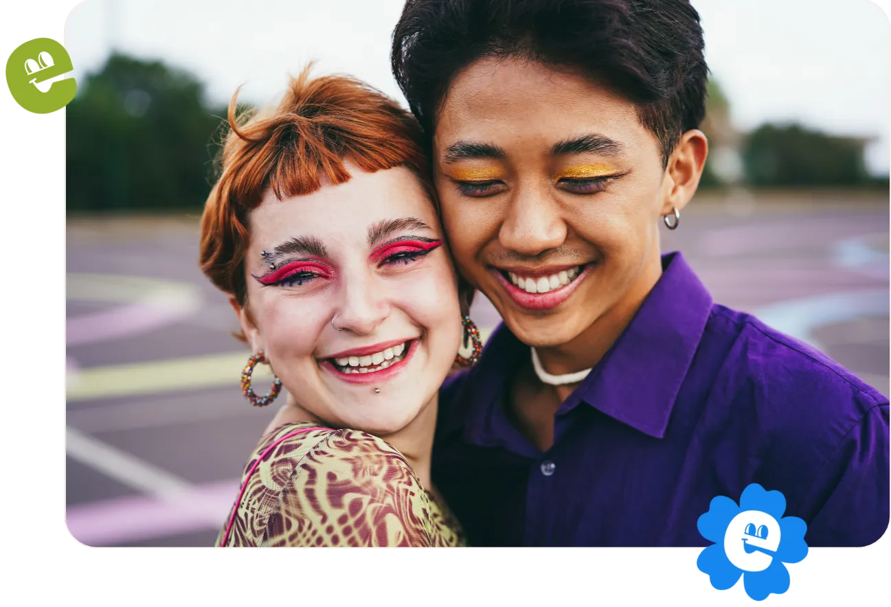 Two smiling young LGBTQ friends standing cheek to cheek. They are both wearing colorful eye make up.