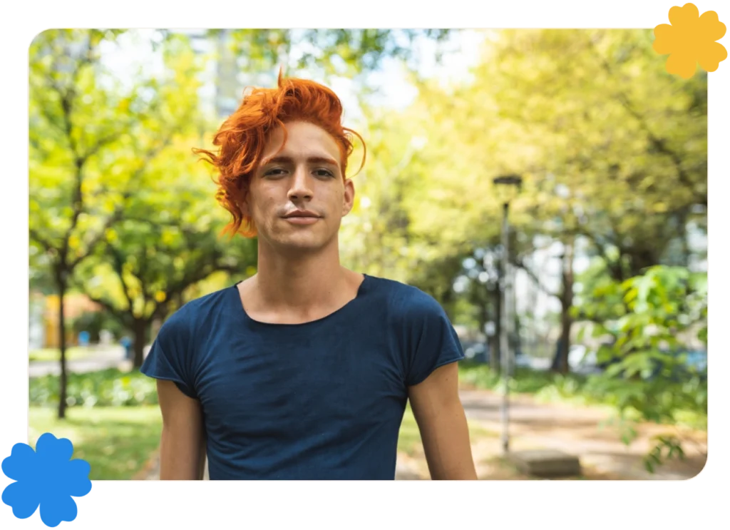 A young LGBTQ person with died red hair stands confidently in front of an urban park.