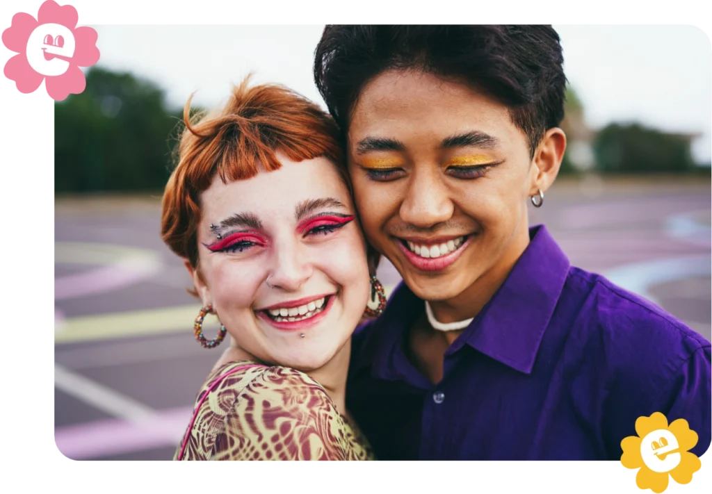 Two smiling young LGBTQ friends standing cheek to cheek. They are both wearing colorful eye make up.