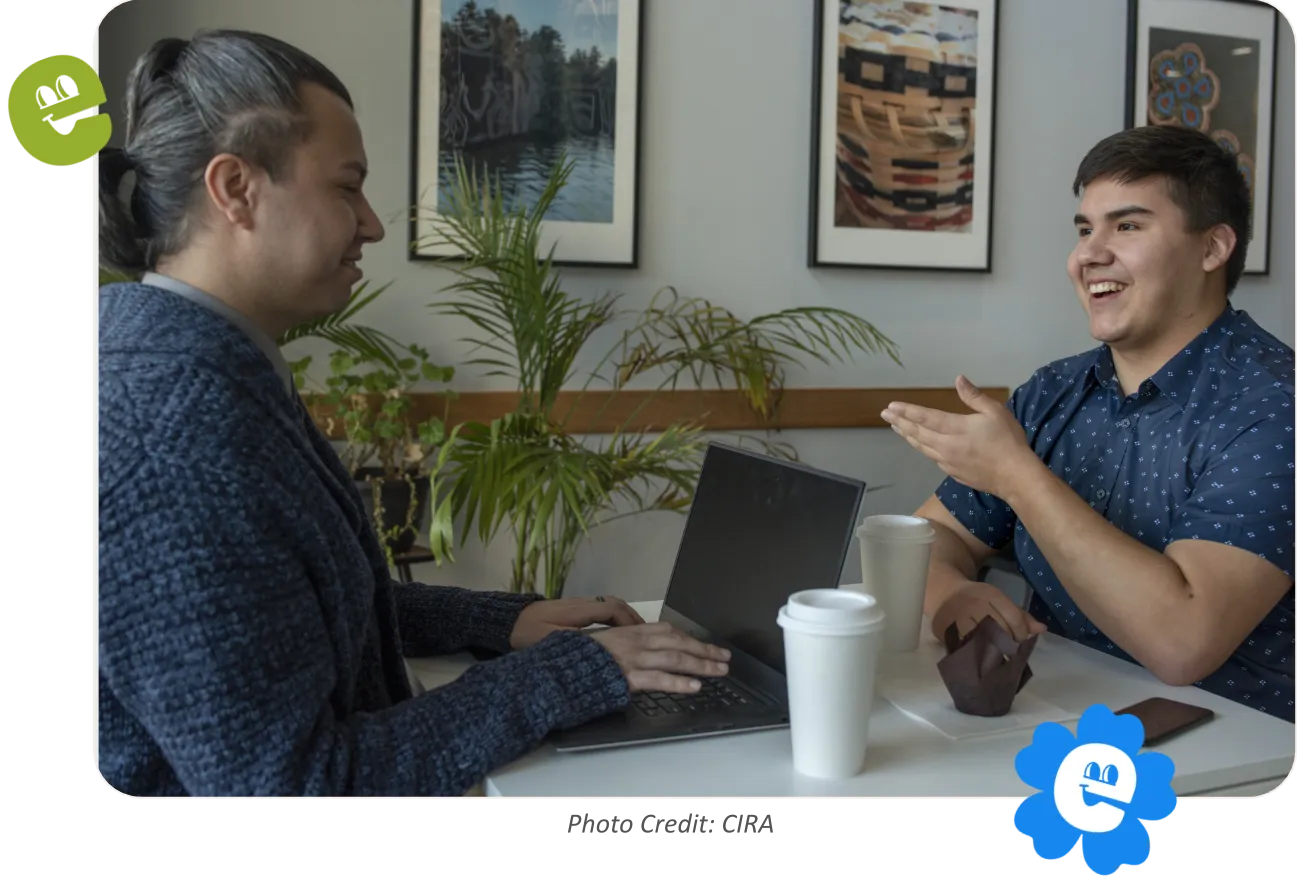 A smiling young First Nations man sits at a table across from an older First Nations man. Indigenous artwork is displayed on the wall behind them.
