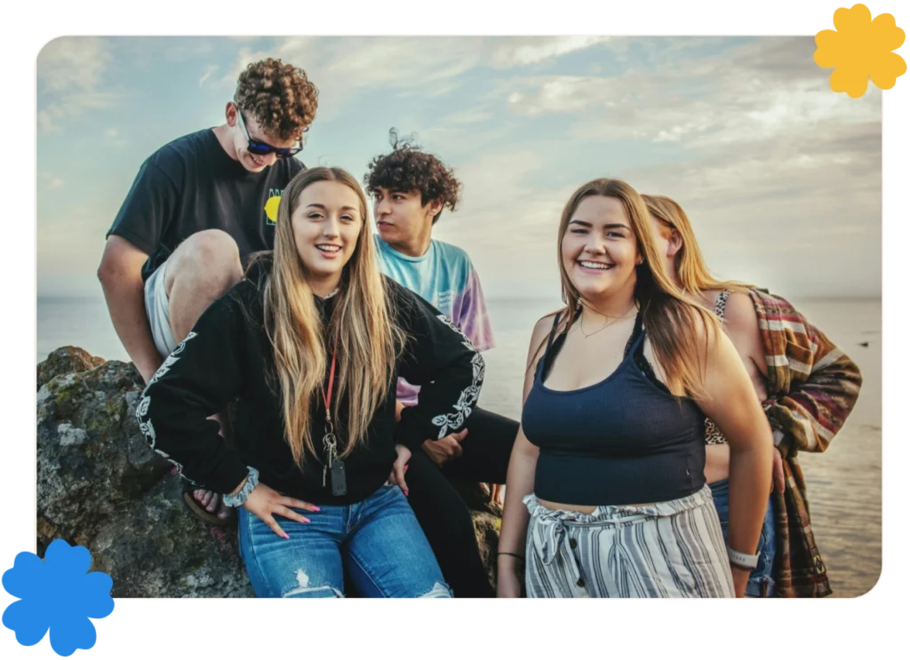 A group of youth sit on a rock in front of the ocean. Two girls in front smile at the camera.